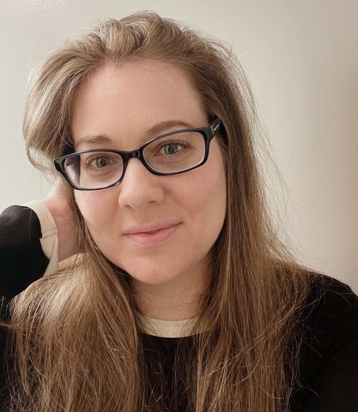 Headshot: young white woman with long light-brown hair and black glasses, soft smile, hand at temple, neutral light background.